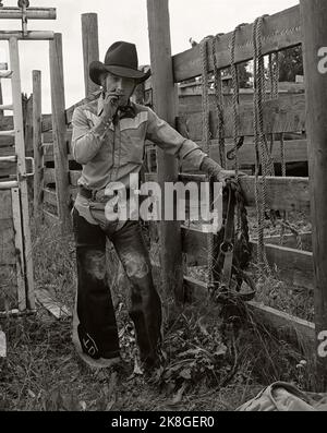 Monochromes Vintage-Porträt eines Rodeo-Cowboys mit Lederbeinen und Lehnen an einer Rutsche in einer Schrift über Stone, Alberta, Kanada. Um 1983 Stockfoto