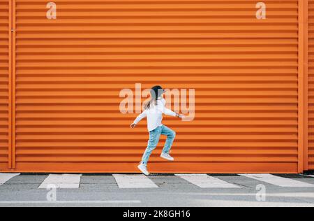 Kleine Mode-gekleidete Mädchen lustig springen über einen Fußgängerzebra Crosswalk auf der orangen Wand Hintergrund. Urbane Menschen leben und eine Straße jeden Stockfoto