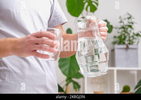 Ein Mann gießt kaltes Wasser in ein Glas. Nahaufnahme von männlichen Händen, die Wasser aus einem Krug in einen Glasbecher gießen. Ein Mann löscht seinen Durst. Stockfoto