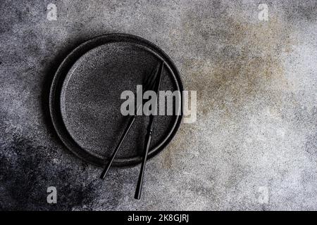 Von oben minimalistische Tischunterlage mit dunklem Steinzeug und schwarzem Besteck auf Betonhintergrund Stockfoto