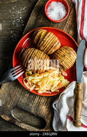 Gebackene Hasselback-Kartoffeln mit Gewürzen und fermentierter Kohlsalat in einer Schüssel auf einem Holztisch serviert Stockfoto