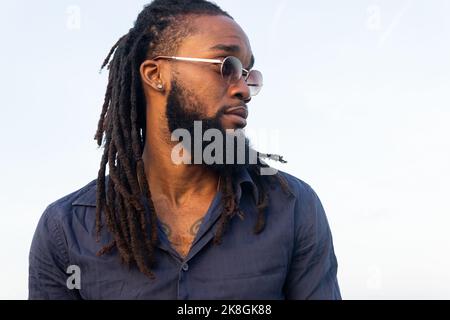 Schwarzer Rüde mit Dreadlocks in Sonnenbrillen, der an der Küste steht und gegen den grauen Himmel blickt Stockfoto