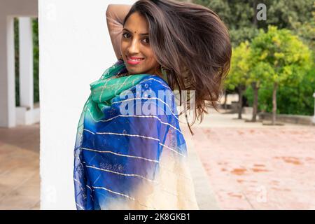 Positive junge indische Frau in traditioneller Kleidung und Schal lächelnd und Blick auf die Kamera im grünen Park Stockfoto