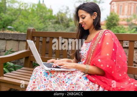 Seitenansicht einer jungen indischen Freiberuflerin in Kleidung, die auf einer Holzbank sitzt und auf einem Laptop tippt, während sie an einem Remote-Projekt arbeitet Stockfoto