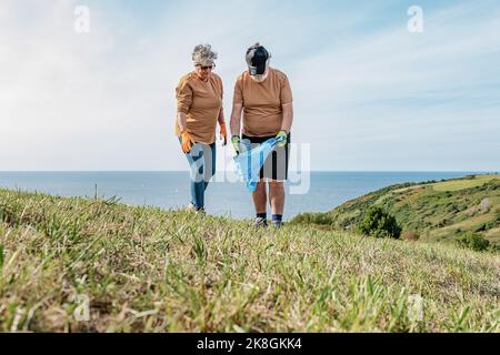 Ältere Freiwillige und Freiwillige in Uniformen sammeln Müll in Plastiktüten auf einem grasbewachsenen Hügel in der Nähe des Meeres gegen den Himmel Stockfoto