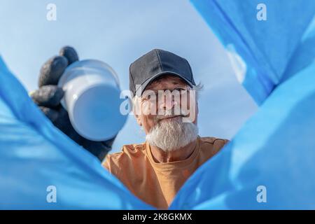 Von unten aus wurden bei der Müllabfuhr positiv gealterte bärtige männliche Freiwillige mit Mütze und Schutzhandschuhen in den Müllbeutel geworfen Stockfoto