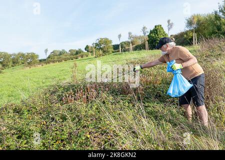 Seitenansicht ganzer Körper eines älteren männlichen Freiwilligen in Mütze, der auf einer Wiese mit Müllbeutel steht und an sonnigen Tagen Müll über eine Mülltonne sammelt Stockfoto