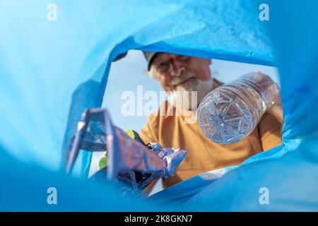 Von unten aus wurden bei der Müllabfuhr positiv gealterte bärtige männliche Freiwillige mit Mütze und Schutzhandschuhen in den Müllbeutel geworfen Stockfoto