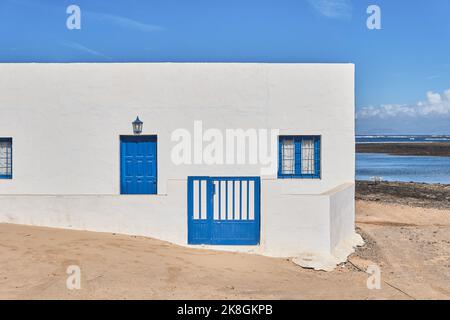 Außenansicht eines typischen weißen kanarischen Wohnhauses mit blauer Tür und Fenstern an einer sandigen Küste in der Nähe des Ozeans gegen den wolkigen blauen Himmel an sonnigen Tagen in Fuer Stockfoto