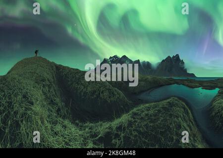 Nicht erkennbarer Fotograf mit Fotokamera, die auf einer Bergklippe steht und den Blick auf die grüne aurora Borealis einfängt, die den dunklen Nachthimmel erhellt Stockfoto