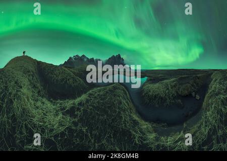 Nicht erkennbarer Fotograf mit Fotokamera, die auf einer Bergklippe steht und den Blick auf die grüne aurora Borealis einfängt, die den dunklen Nachthimmel erhellt Stockfoto