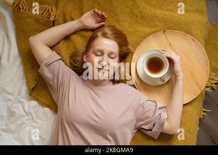 Draufsicht auf eine positive junge rothaarige Dame, die auf einem karierten Kleid mit geschlossenen Augen neben einer Tasse heißen Tee auf einem runden Holztablett lag Stockfoto