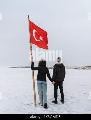 Rückansicht eines anonymen jungen Paares in warmer Kleidung, das die Hände hielt, während es auf verschneitem Gelände mit einer großen türkischen Flagge gegen den bewölkten Himmel stand Stockfoto