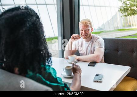 Eine nicht erkennbare afroamerikanische Frau mit einem fröhlichen blonden Mann im weißen T-Shirt, der mit Tassen Kaffee am Tisch sitzt und sich jede Person ansieht Stockfoto