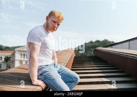 Die Seitenansicht des jungen bärtigen, muskulösen blonden Mannes in weißem T-Shirt und Jeans, der auf einem Handlauf einer Holztreppe gegen die verschwommene Stadt sitzt und anschaut, kam Stockfoto