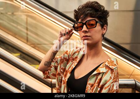 Junge Brünette mit kurzen Haaren, die die Kamera anschaut und eine stilvolle Sonnenbrille anpasst, während sie in der Nähe einer unterirdischen, beweglichen Treppe in Madrid, Spanien, steht Stockfoto