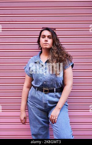 Serious junge Brünette in Denim-Kleidung Blick auf die Kamera, während in der Nähe rosa Wand des modernen Gebäudes auf der Straße von Madrid, Spanien stehen Stockfoto