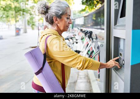 Crop Senior weiblich mit Smartphone, um Zugang zu ATM in der Nähe von Elektro-Fahrrad-Verleih-Station auf der Straße der Stadt zu erhalten Stockfoto