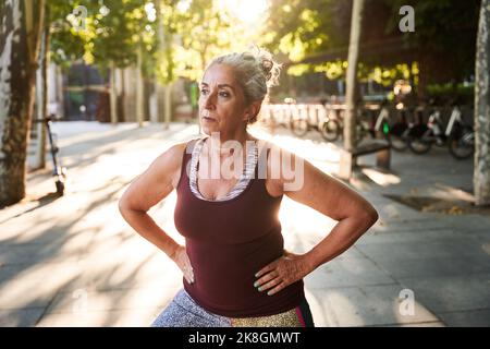 Gealterte Frau mit sportlicher Oberseite und Leggings, die während des Fitnesstrainings auf dem gepflasterten Bürgersteig in der Nähe des Parks an sonnigen Tagen Ausfallschritte mit den Händen auf der Hüfte macht, während sie sich auf der Toilette aufhält Stockfoto
