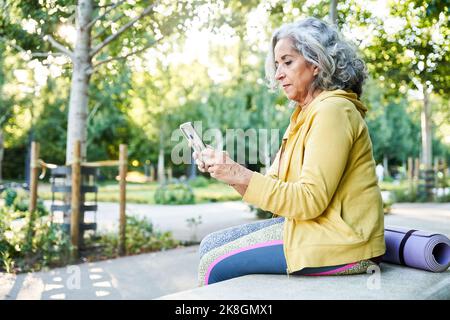 Ganzkörper-Seitenansicht der konzentrierten Senior Dame mit kurzen grauen Haaren in Sportkleidung sitzen auf Steinbank in der Nähe gerollte Yoga-Matte und Surfen im Internet V Stockfoto