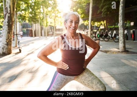 Eine fröhliche, gealterte Frau in sportlichem Oberteil und Leggings, die während des Fitnesstrainings auf dem gepflasterten Bürgersteig in der Nähe des Parks an sonnigen Tagen Ausfallschritte mit den Händen auf der Hüfte macht Stockfoto