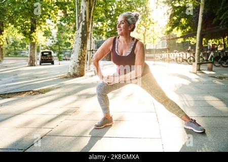 Eine fröhliche Frau im Alter in sportlichem Oberteil und Leggings, die während des Fitnesstrainings auf dem gepflasterten Gehweg in der Nähe des Parks an sonnigen Tagen die Beine streckt, während sie schaut Stockfoto