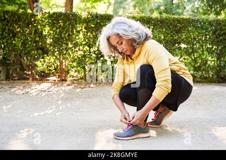 Die ältere Sportlerin kniet auf dem Bürgersteig und bindet Schnürsenkel auf den Sneaker, während sie sich früh am Morgen in der Stadt auf das Fitnesstraining vorbereitet Stockfoto