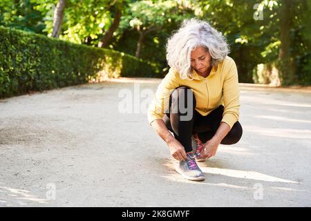 Die ältere Sportlerin kniet auf dem Bürgersteig und bindet Schnürsenkel auf den Sneaker, während sie sich früh am Morgen in der Stadt auf das Fitnesstraining vorbereitet Stockfoto