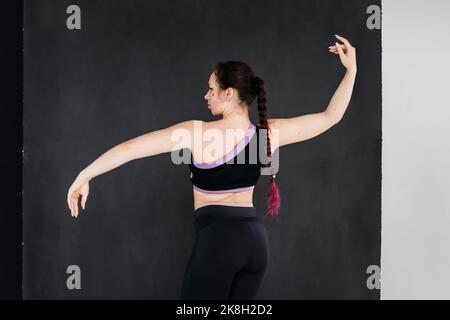 Eine hübsche Brünette Mädchen tanzen. Dunkler Hintergrund. Sanfte Körperbewegungen. Studioportrait einer jungen Sportlerin in einer schwarzen Hose und einem Oberteil. Weiblichkeit Co Stockfoto