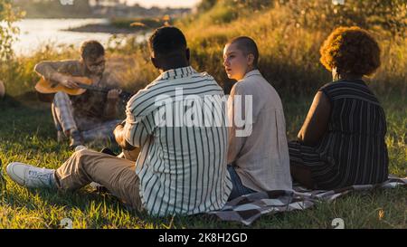 Im Sommer oder Herbst am Fluss. Drei multirassische Freunde sitzen auf karierter Picknickdecke und nehmen. Verschwommener kaukasischer Typ mit akustischer Gitarre im Hintergrund. Hochwertige Fotos Stockfoto