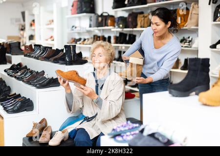 Freundliche weibliche Verkaufsberaterin bedient eine reife Frau, die sich Schuhe anprobieren wird Stockfoto