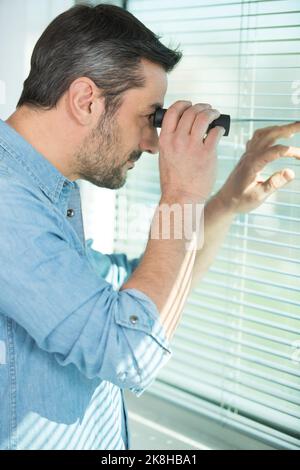 Mann, der durch ein Fernglas auf das Fensterblind blickt Stockfoto