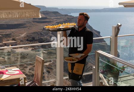 Fira, Santorini, Griechenland. 2022. Kellner, der Essensgerichte auf einem Bistro auf einer Klippe in Fira, Santorini, mit Blick auf die Ägäis, Santorini, trägt. Stockfoto