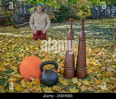 Schwere eiserne Kugelhantel, Slam Ball und hölzerne persische Keulen in einem Hinterhof mit einem älteren männlichen Seiza, der in einer Hintergrund-Herbstlandschaft sitzt Stockfoto