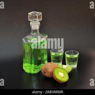 Likör auf Kiwi-Basis, hausgemachter Alkohol in einer Flasche und zwei Gläser auf schwarzem Hintergrund, neben reifen geschnittenen Früchten. Nahaufnahme. Stockfoto