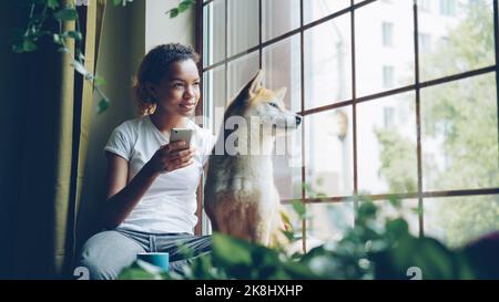 Die hübsche junge Frau sitzt auf der Fensterbank und nutzt ihr Smartphone, während ihr netter, ruhiger shiba-Inu-Hund neben ihrem Blick sitzt. Freizeit, Tiere und Häuser Konzept. Stockfoto