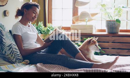 Attraktives lächelndes Mädchen liest Buch und streichelt schönen Hund auf dem Bett in der Nähe seines Besitzers zu Hause liegen. Modernes Interieur mit großen Fenstern, grünen Pflanzen und Holzdekor ist sichtbar. Stockfoto