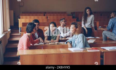 Eine Gruppe von Universitätsstudenten mit mehreren Rassen entspannen sich und plaudern in der Pause und genießen Freizeit und Kommunikation. Holztische, attraktive Menschen und Notizbücher sind sichtbar. Stockfoto