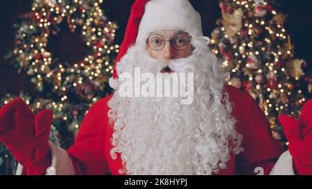 Porträt eines Mannes in traditioneller Weihnachtsmann-Tracht und Brille beim Blick auf die Kamera in der Nähe des Weihnachtsbaums in einem dunkel beleuchteten und dekorierten Raum. Urlaub und Menschen Konzept. Stockfoto
