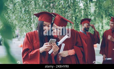 Weibliche Absolventen nutzen Smartphone, um auf dem Bildschirm zu reden und zu lachen, stehen im Freien mit Diplomen, hübsche Mädchen tragen formelle Kleider und Hüte. Stockfoto