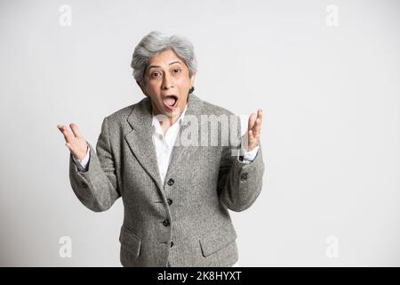 Reife indische Senior Geschäftsfrau Chef in Anzug stehend mit ihrem Mund offen in überrascht oder schockiert vor weißem Studio Hintergrund, Asian 60s grau-h Stockfoto