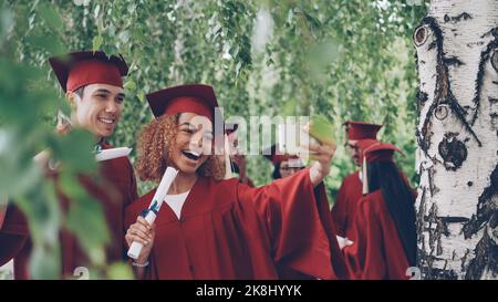 Glückliche junge Menschen Mädchen und Kerl nehmen Selfie nach der Abschlussfeier mit Diplomen tragen Kleider und Mortarboards. Fotografien, Jugend- und Bildungskonzept. Stockfoto