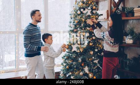 Eltern und Kind schmücken den Neujahrsbaum in einer modernen Wohnung und unterhalten sich miteinander. Schönes Interieur, Menschen und Urlaub Konzept. Stockfoto
