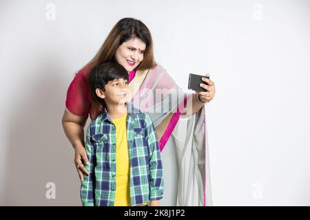 Porträt einer glücklichen indischen Mutter und eines Sohnes mit einem Smartphone, das Selfie macht, Videoanruf, isoliert auf weißem Studiohintergrund, Frau im Sari-Stehen Stockfoto