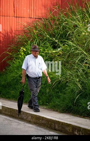 Filandia, Quindio, Kolumbien - Juni 5 2022: Alter Mann mit Hut und Brille geht mit einem schwarzen Regenschirm den Bürgersteig hinunter Stockfoto