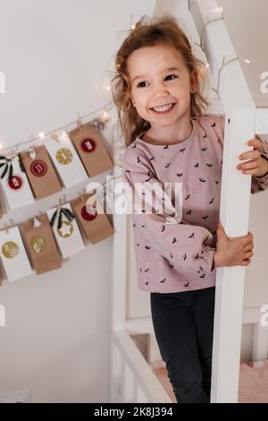 Kleinkind Kind bekommt neue Adventskalender Aufgabe, warten auf Weihnachten. Festliche Stimmung für niedliche lockige Mädchen. Kid nimmt Adventsbeutel mit Daten von der Wand an Stockfoto