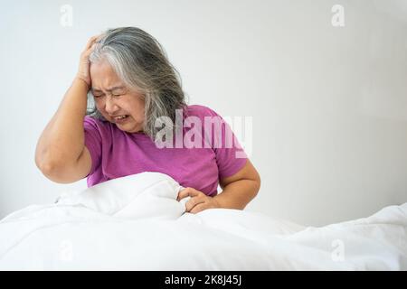 Ungesunde ältere asiatische Senior-Frau auf dem Bett sitzen unter Kopfschmerzen und Migräne so Schmerzen und Krankheit, Stress ältere Frau sitzen auf dem Bett zu Hause Stockfoto