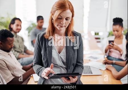 Ein weiterer Weg zum Erfolg: Eine reife Geschäftsfrau, die in einem Büro ein digitales Tablet nutzt, mit ihren Kollegen im Hintergrund. Stockfoto