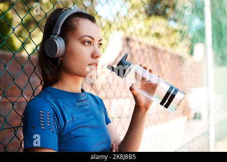 Eine sportliche junge Frau trinkt Wasser, während sie im Freien gegen einen Zaun steht. Stockfoto