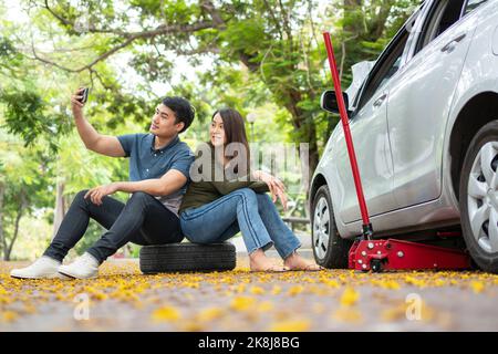 Asiatisches Paar mit Smartphone für Hilfe nach einem Auto Panne auf der Straße. Konzept des Motorproblems des Fahrzeugs oder der Unfall- und Notfallhilfe von PR Stockfoto